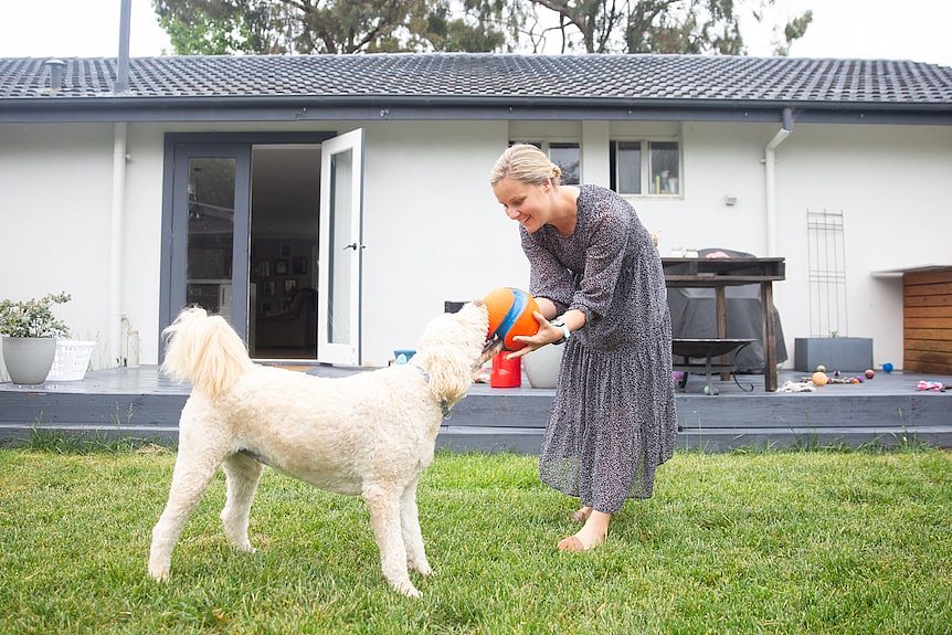 A woman in a dress plays with a white dog and a ball in a backyard.