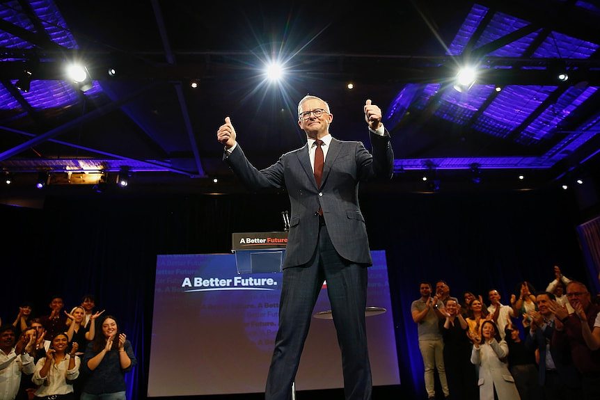 Albanese gives a thumbs up from a stage as an audience applauds him, with campaign slogans visible behind him.