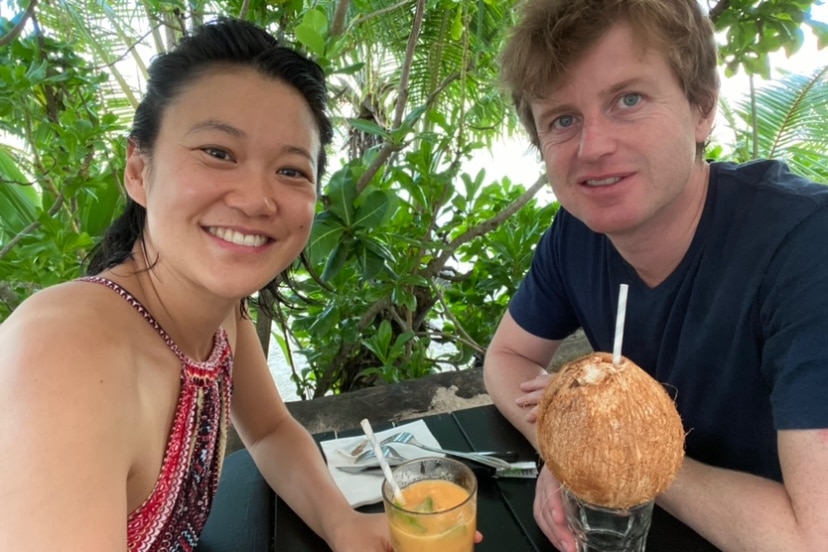 An asian woman smiling with a white male drinking out of coconuts with green background 