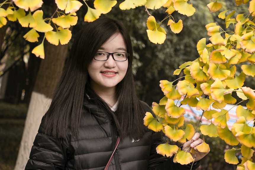 An Asian girl wearing a black puffer jacket and black glasses smiling with bright yellow flowers. 