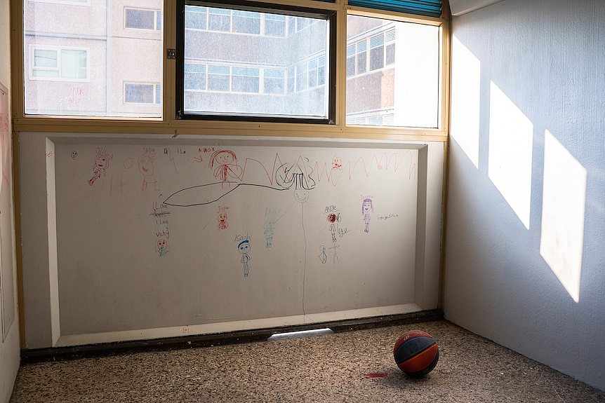 A child's drawings mark an apartment wall beneath a window. A basketball sits on the bare floor.