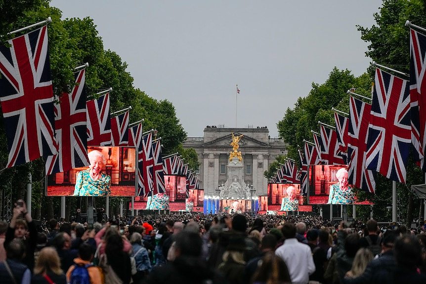 A huge crowd in front of Buckingham Palace, the road lined with British flags hung and The Queen on screens