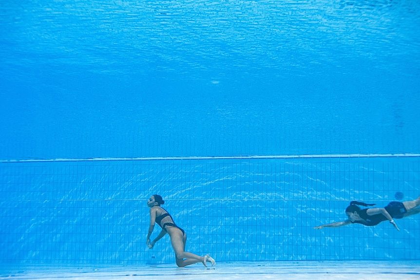 A woman swims towards another woman who is unconscious at the bottom of a pool.