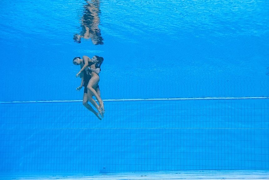 A woman, carrying an unconscious woman, swims from the bottom of a pool to the surface.