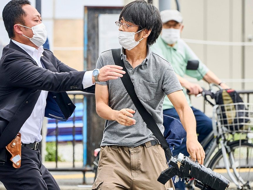 Masked man in grey shirt holding a crude firearm is apprehended by suited security personnel.