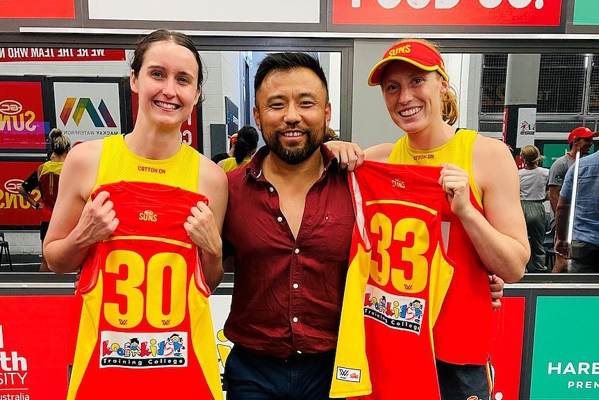 An Asian man taking photos with two AFLW players.