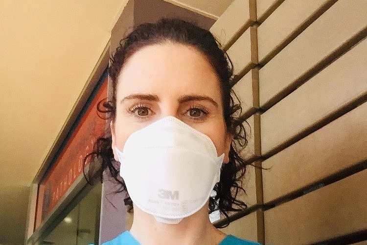 A close up profile image of Kerry with dark curly short hair, hazel eyes wearing white surgical mask infront of wall 