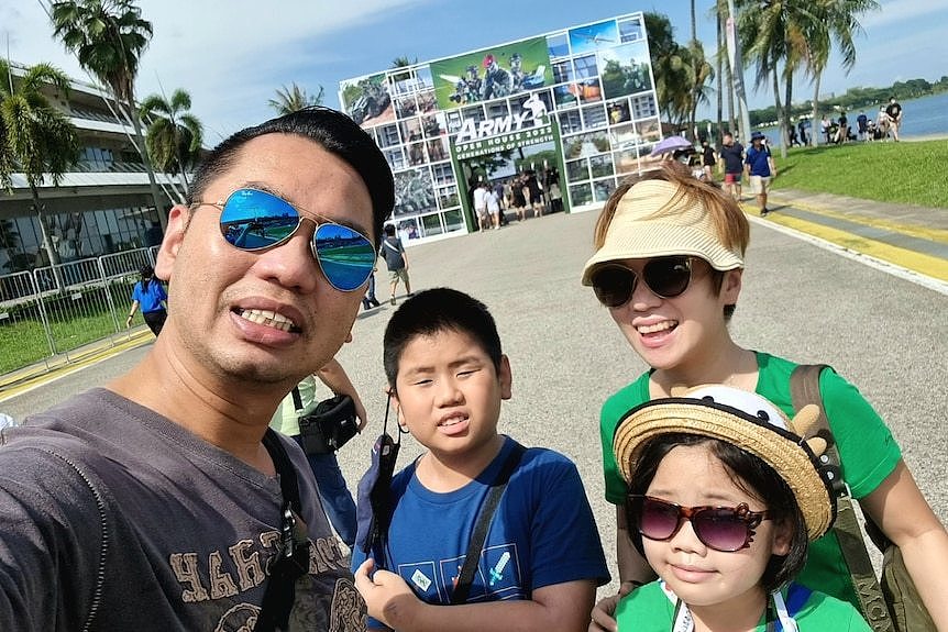 An Asian man and his Asian wife wearing sunglasses pose for selfie with a boy and girl in tropical city setting