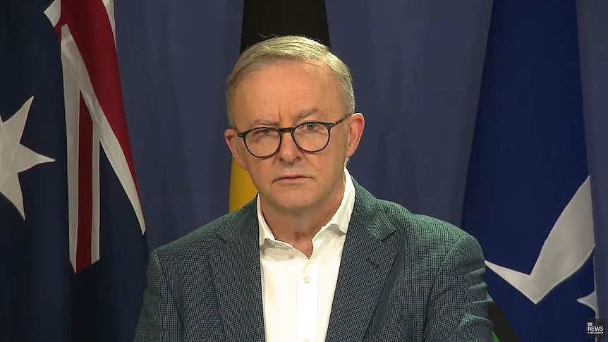 Anthony Albanese in a suit and glasses in front of australian, indigenous and torres strait island flags