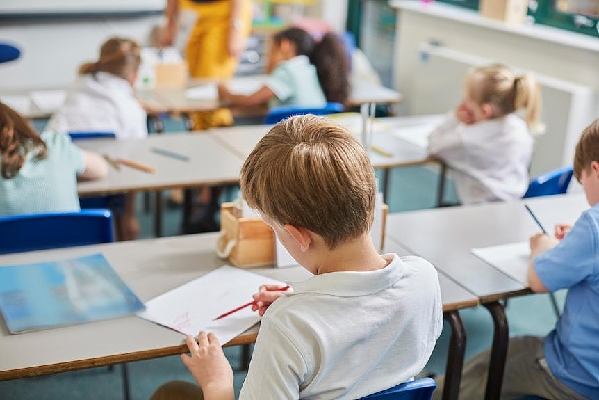 The back of a boy with blonde hair sitting at a school desk going over his work. 