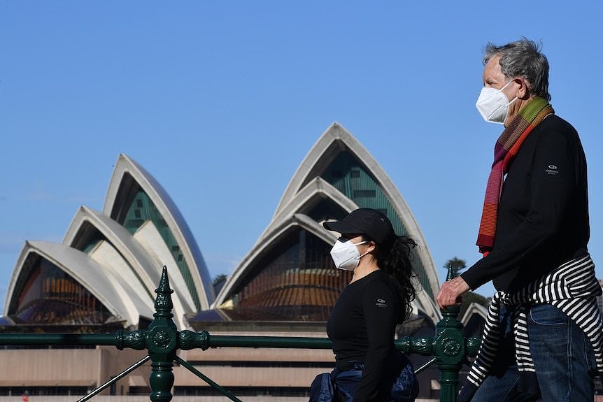 A man and a woman wearing masks walk past the Sydney Opera House. 