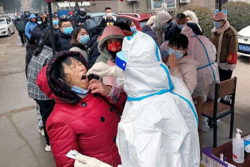 A woman closes her eyes as a person in full PPE uses a swab to collect material for testing from the woman's mouth.