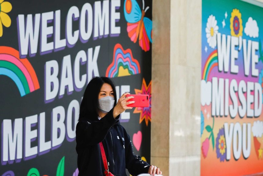 A woman takes a selfie in front of a sign reading 'welcome back Melbourne'.