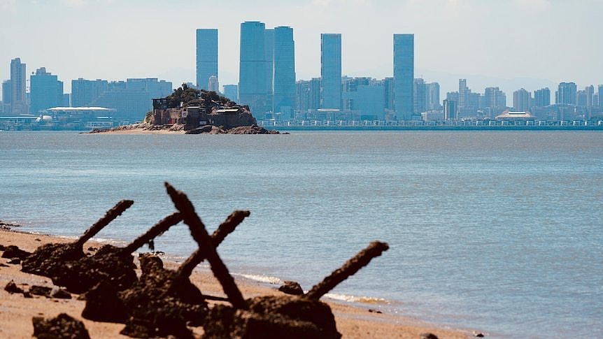 A small island sits in the centre of the sea with the outline of a city behind it.