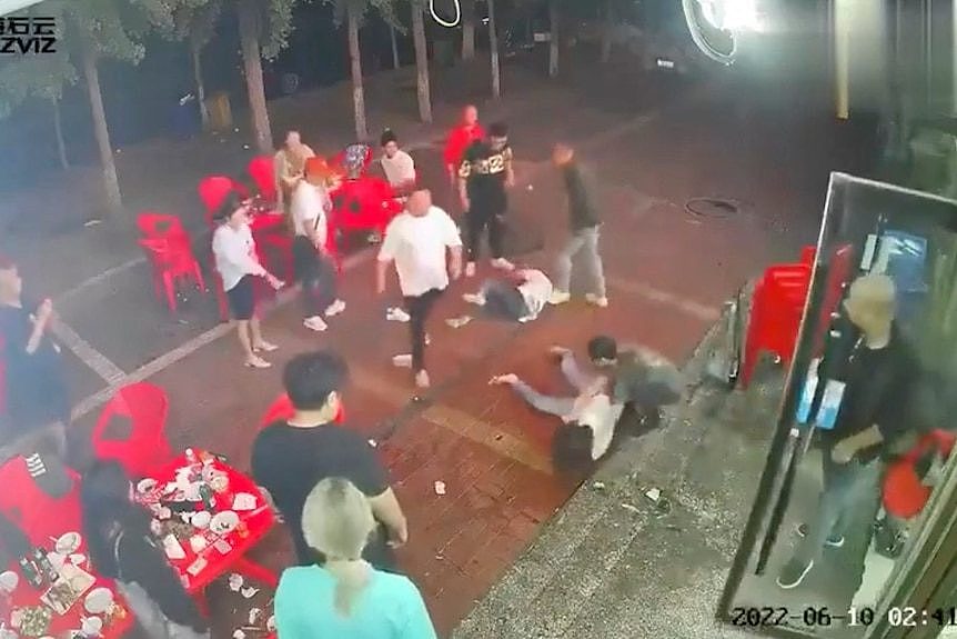 Two women lie on the ground as a group of men surround them outside a restaurant.