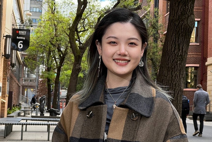 Jinru Sun wears a checked coat and smiles while standing under the trees outside RMIT