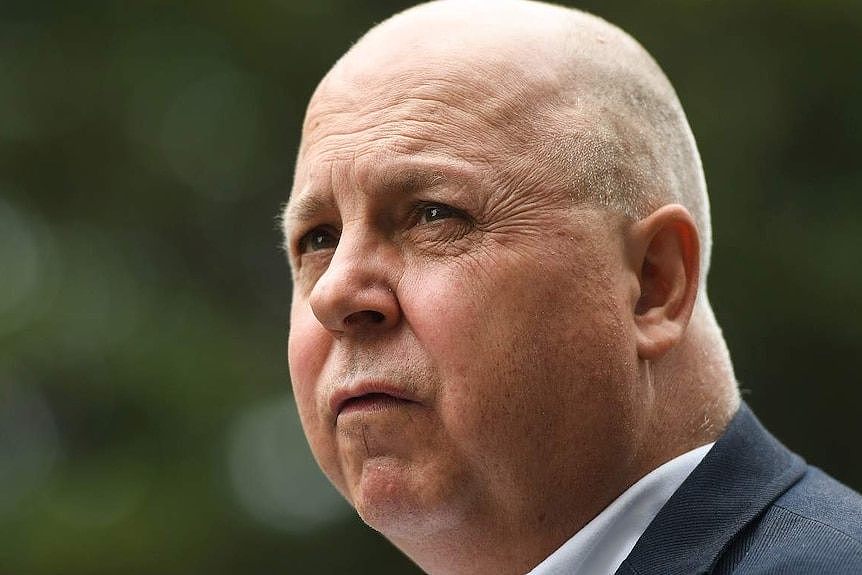 A man with a bald head wearing a suit jacket and shirt stands outside in profile.