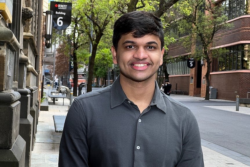 Bhavya wears a grey shirt and stands under the roadside trees outside a university building.