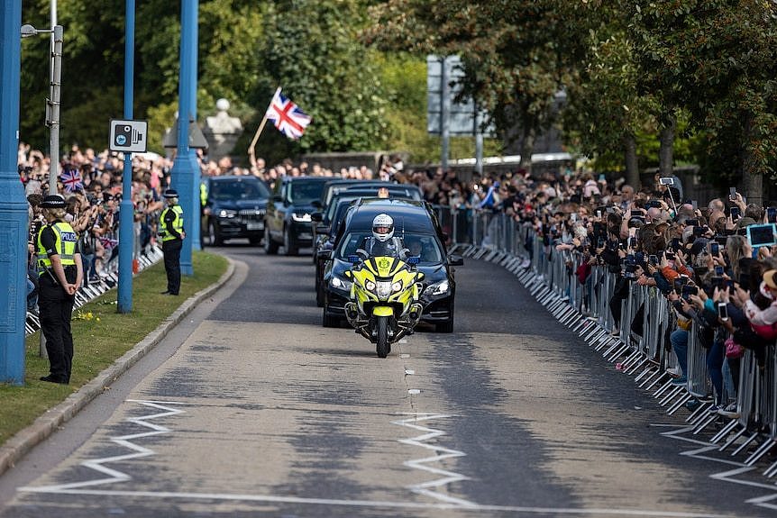 A funeral procession led by a motorbike policeman drives past crowds.