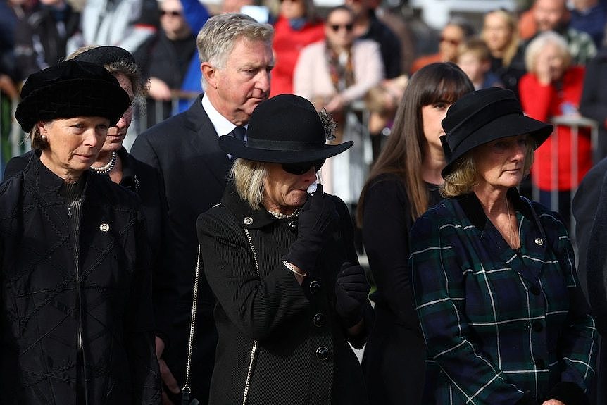 Three women in black clothing appear upset as they watch the hearse pass.