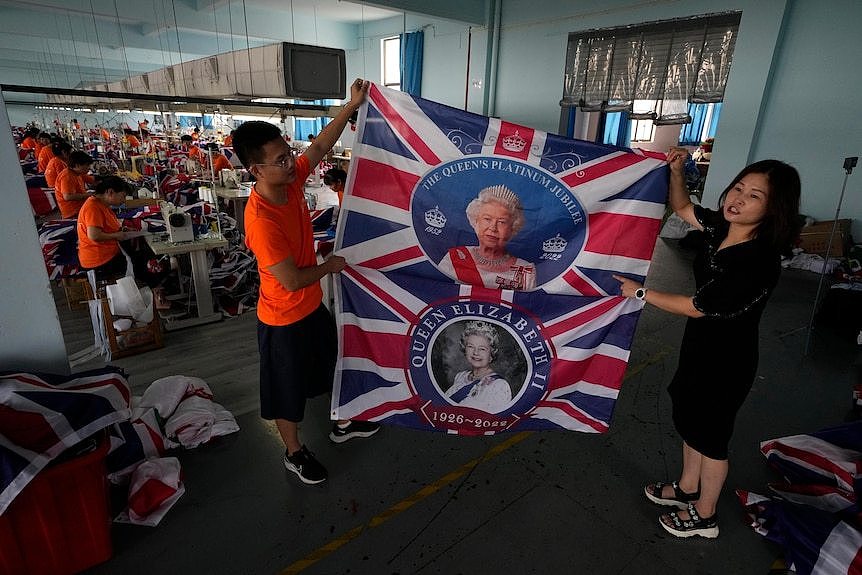On the right a woman and a man are holding up two British flags with an image of the Queen. In the background workers are sowing