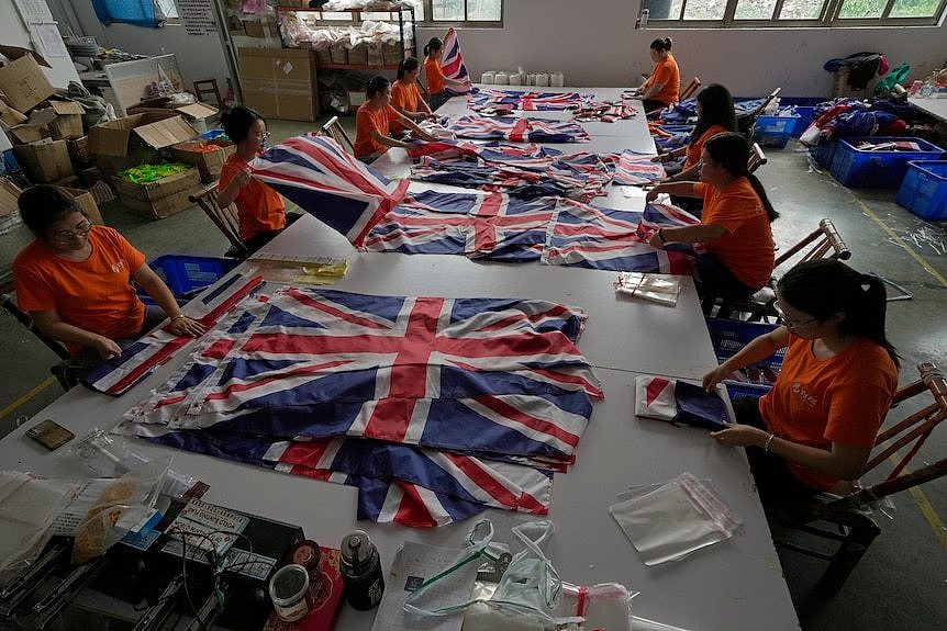 Workers in orange t-shirts are sitting at a large table while folding and packing up British flags.