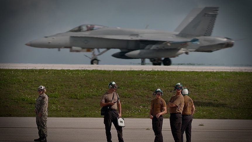 Personnel in khaki Tshirts and camouflage pants stand on a tarmac. Behind them is a fighter jet