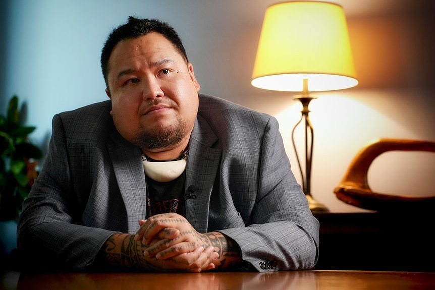 A man wearing a grey checked blazer sits with his tattooed hands clasped together