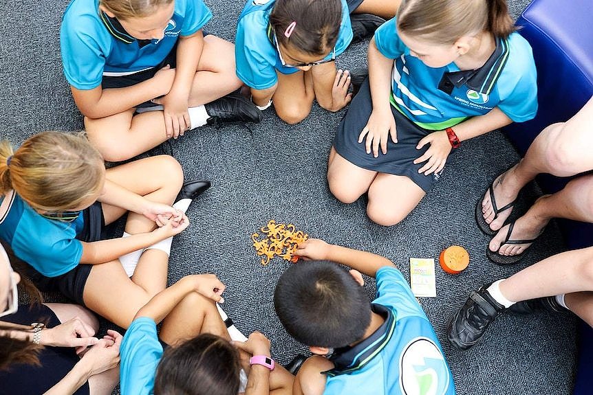 New prep-aged children sit on floor in circle in prep classroom playing barrel of monkeys game.