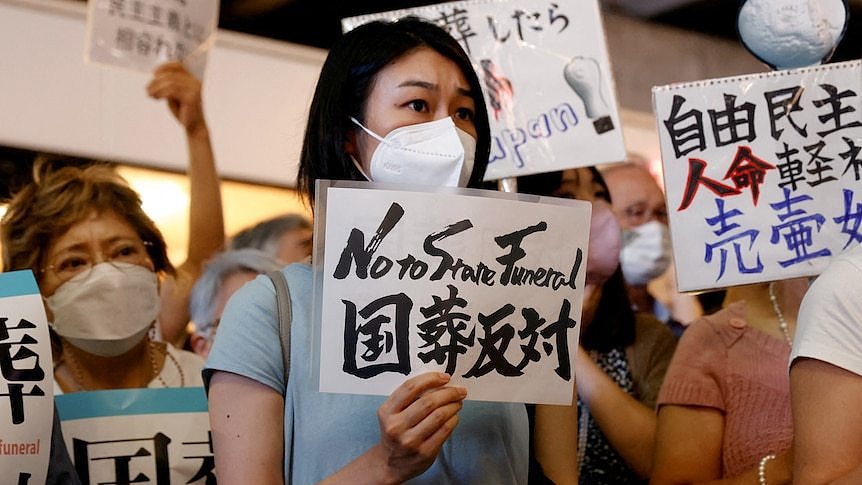 A woman holds a sign saying No to State funeral while wearing a mask surrounded by a crowd of people.