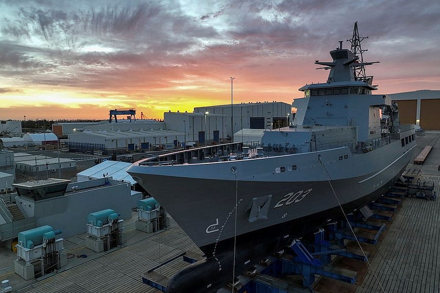 A large naval ship sits on a dock at sunset. 