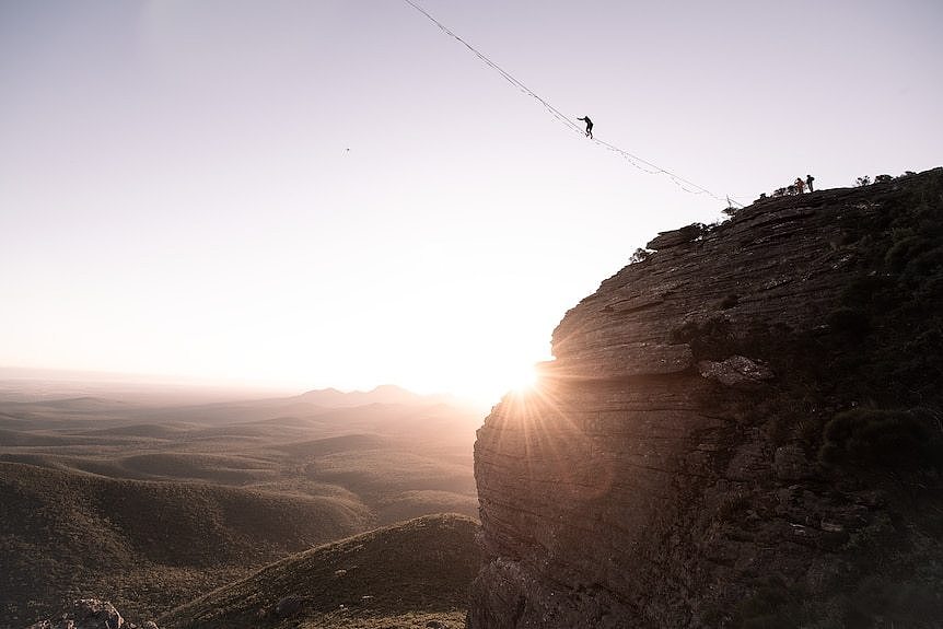 A person suspended on wire as the sun sets behind a rock.