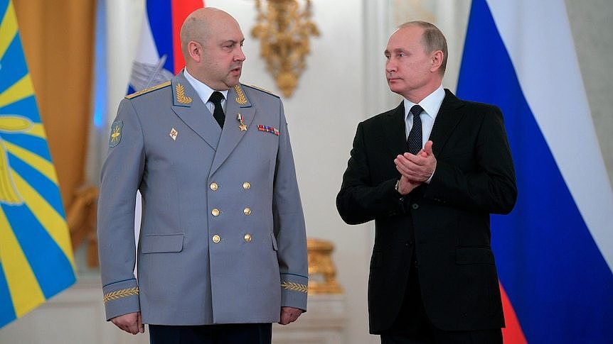Two men stand in front of Russian flags. One is wearing a grey military jacket, the other a black suit. 
