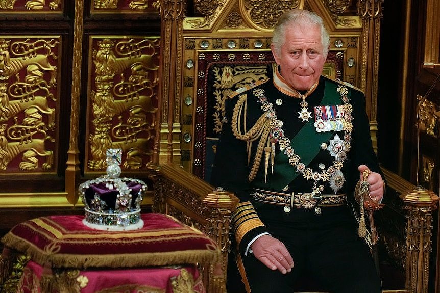 King Charles in full regalia seated next to the Queen's crown