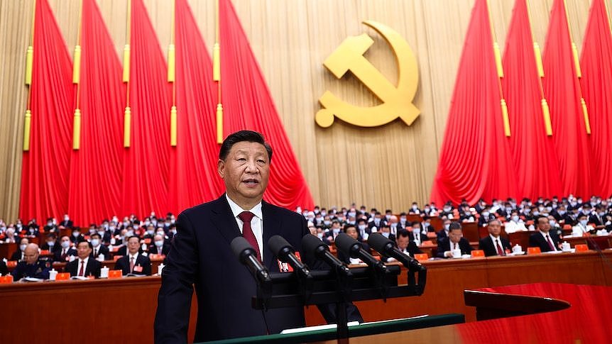A man in a dark suit speaks into a mic in front of a large seated crowd and communist regalia.