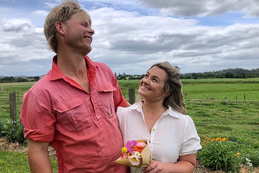 A small woman smiles up at her laughing husband, holding a bouquet of flowers on their farm.