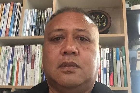 A Samoan man looking blankly at the camera with a bookshelf in the background