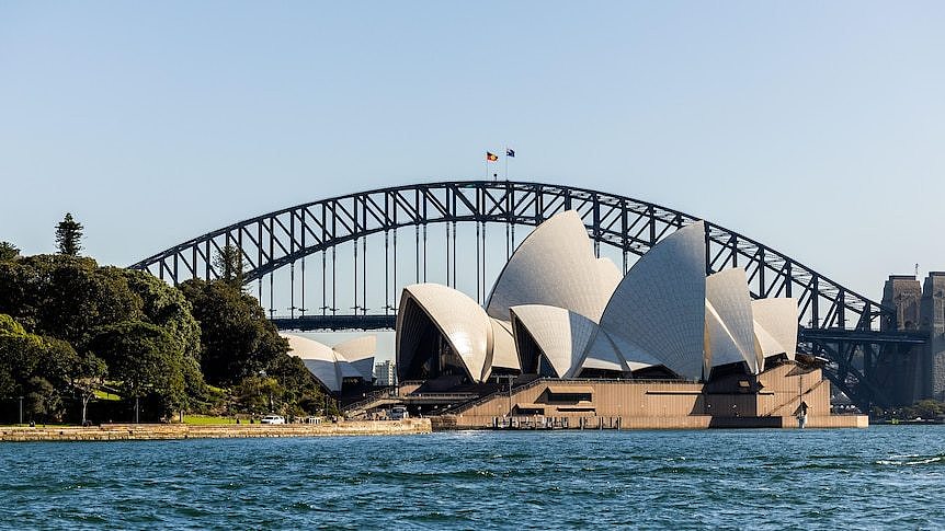 White tiled sails of the Sydney Opera House peak up into a clear blue sky with the Sydney Harbour Bridge in the background.