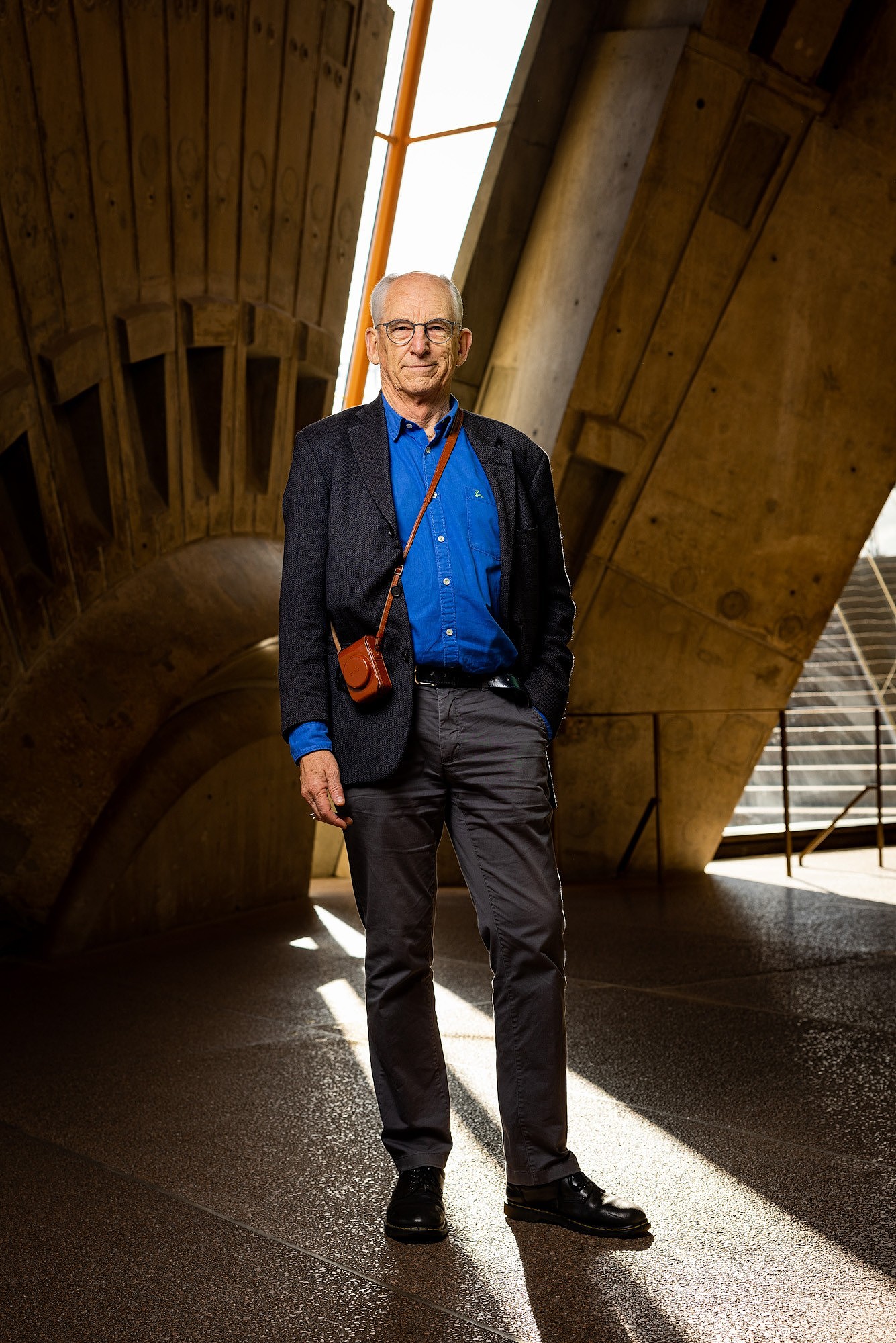 Older white man balding, with white hair and glasses, wearing shirt, chinos and jacket, standing in architectural interior.