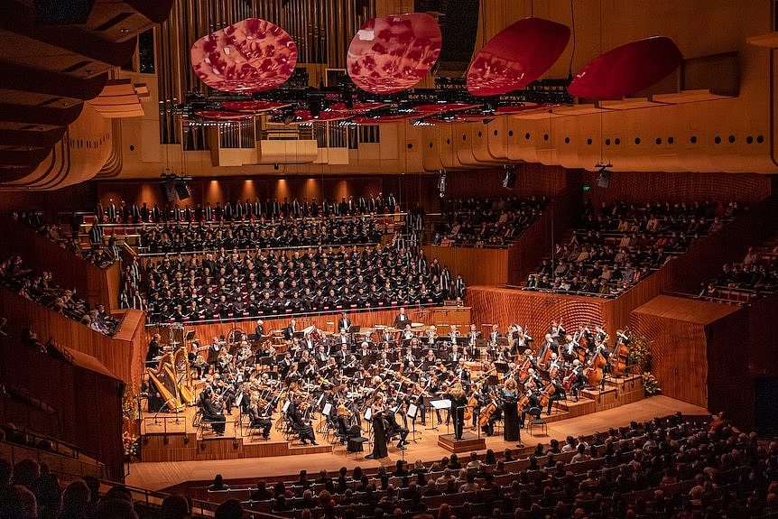 A large orchestra and choir performs on the Sydney Opera House stage. Magenta acoustic reflectors are suspended from ceiling. 