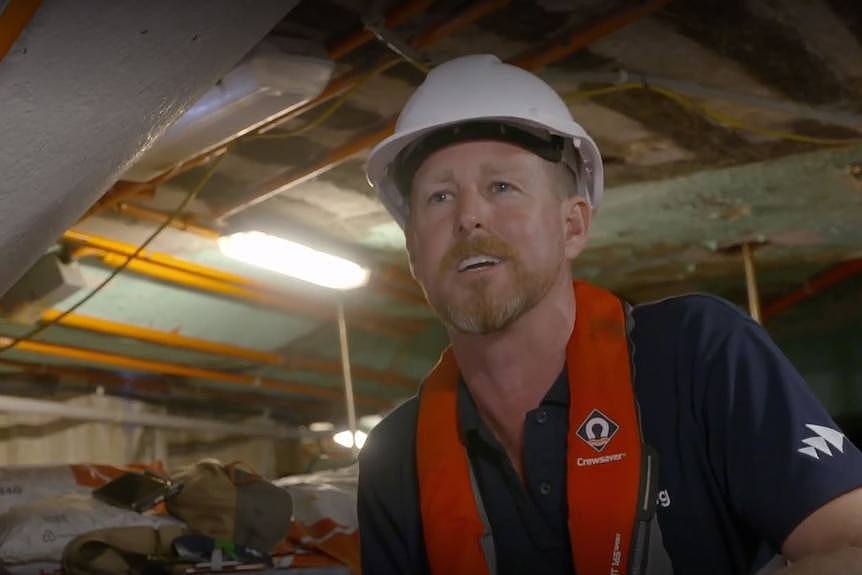 White man wearing construction hard hat and red safety vest speaks to camera from inside an underwater workshop.