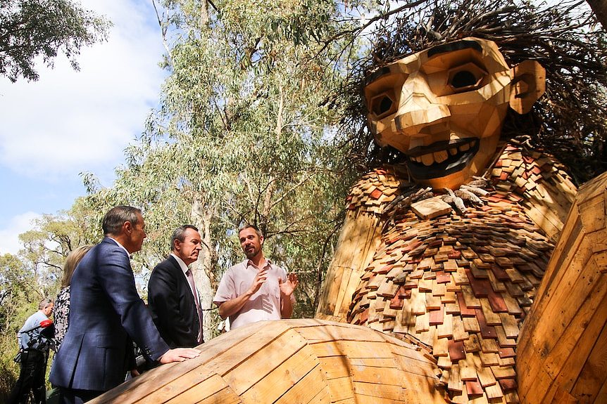 Three men stand next to a huge wooden sculpture