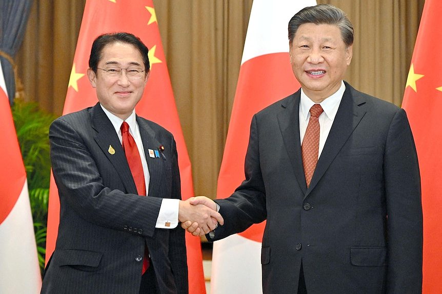 Two men in suits smile as they shake hands in a photo posed before Japanese and Chinese flags.