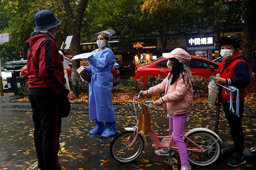 A woman wearing a red jacket holds up her phone to be scanned by a woman dressed in a protective suit.