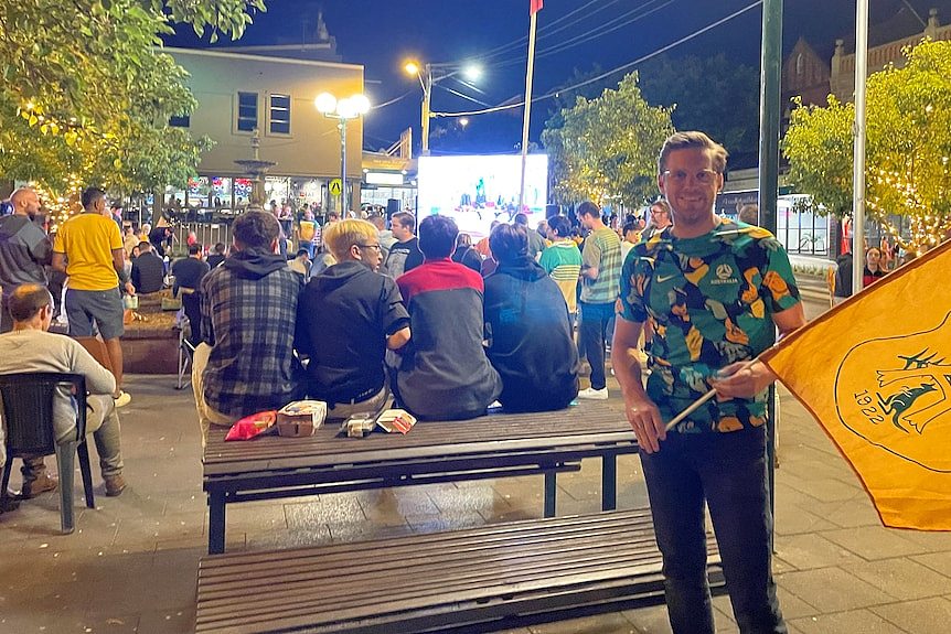 A man holds a yellow flag in front of a crowd of people watching a big screen. 