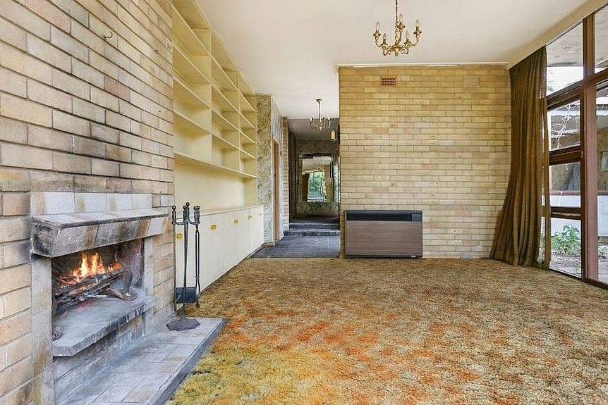 The living room of Gough Whitlam's former home has a fireplace and large bookshelf and floor-to-ceiling windows.