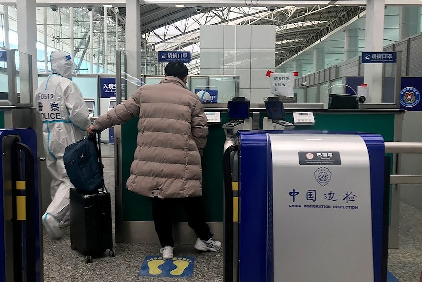 Traveller prepares to cross immigration at airport with police officer in full PPE nearby.