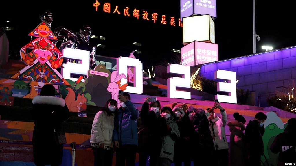 新年前夜，人们站在北京一座大型商场外“2023”字样的灯饰下，而此时新冠病毒正如潮水般在北京卷土重来。霓虹灯庆祝2023年广告背后远处是“中国人民解放军总医院”大楼。