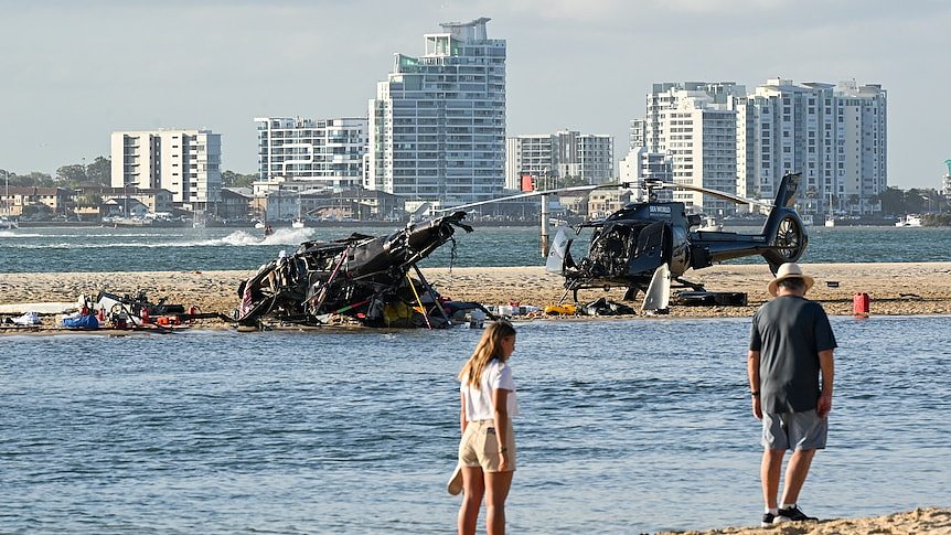 Two people stand on a beach near the wreckage of two helicopters.