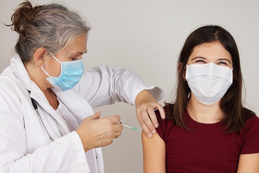 A woman in a white coat and blue face mask vaccinates a teenage girl, who appear to be smiling under her mask.